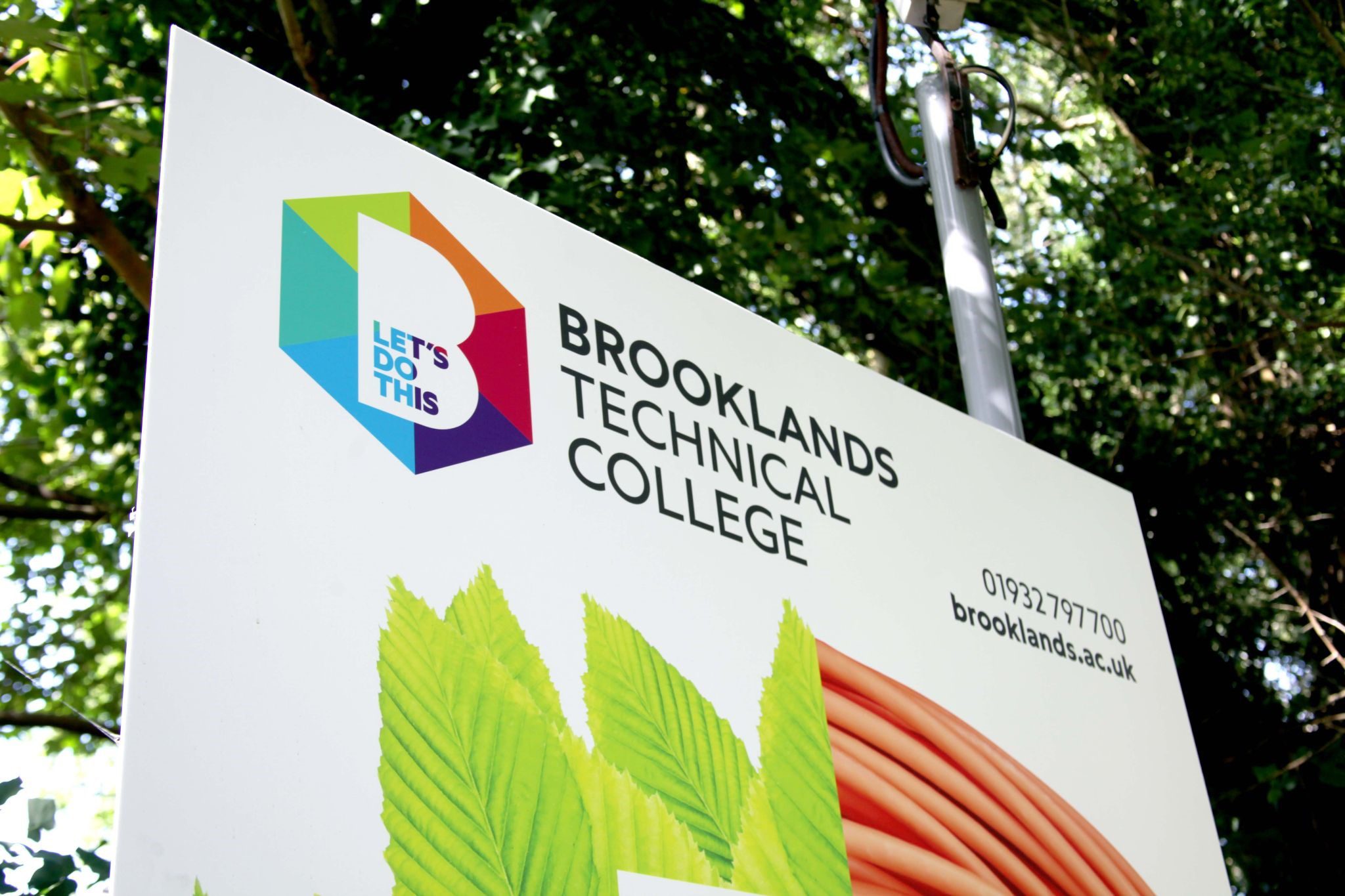 A large white sign depicting the college logo Brooklands Technical College surrounded by trees in the background.